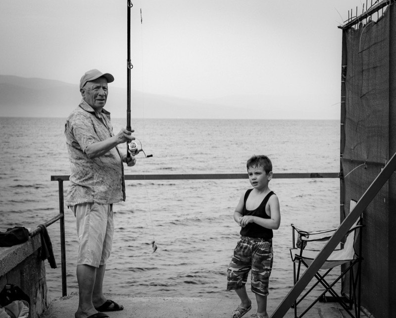 A grandfather fishing from a jetty with a young boy beside him